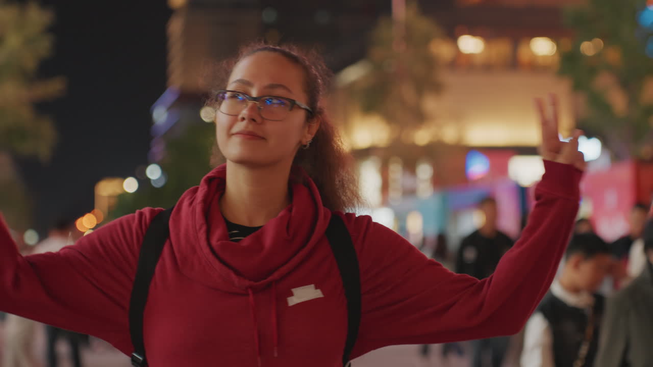 Ritratto stradale notturno di una donna con maglione rosso e occhiali, in pose giocose e che fa il segno della pace; segue una vista posteriore riflessiva mentre si trova in una vivace piazza illuminata, tra luci bokeh soffuse, creando una calda atmosfera di vita notturna urbana.