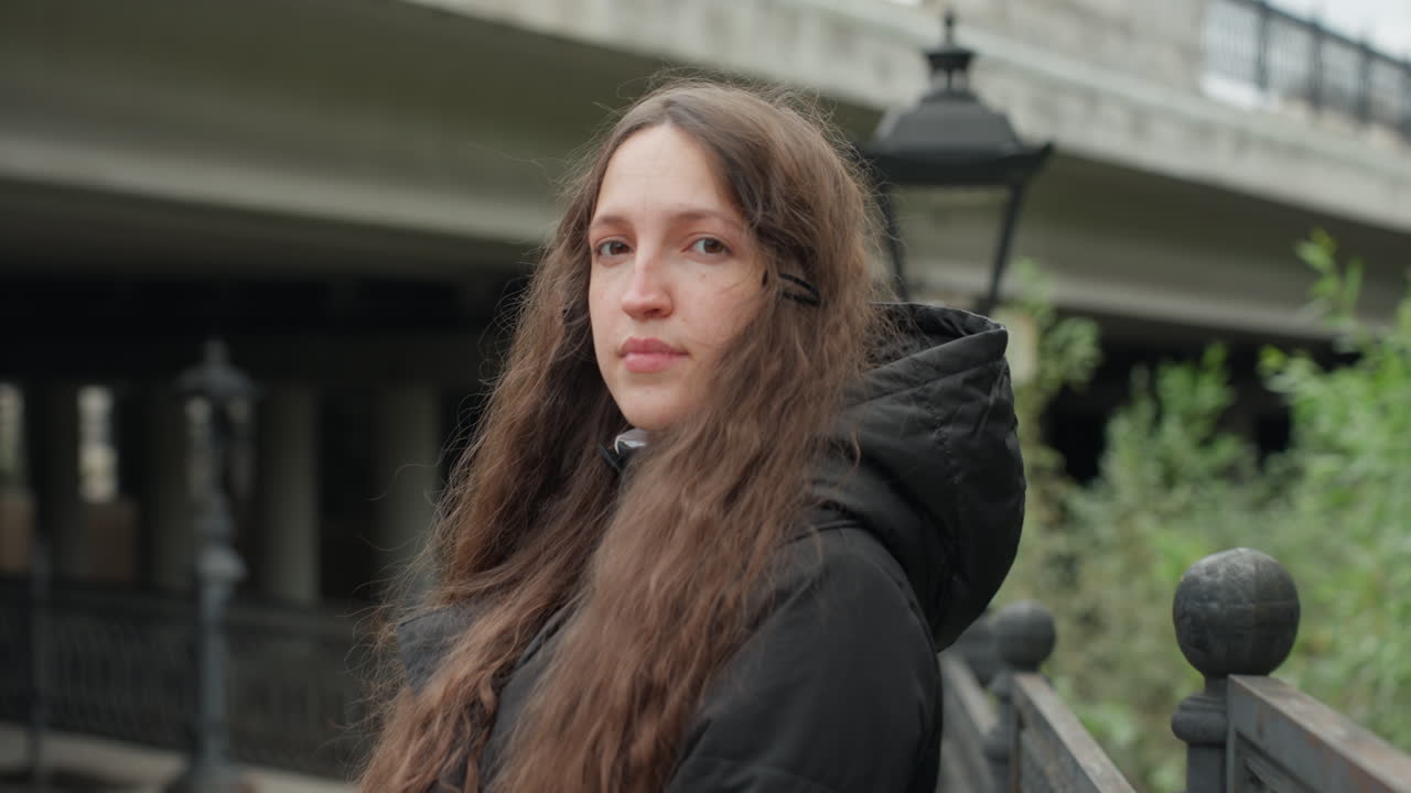 Giovane donna che cammina su un ponte urbano, sequenza di ritratti contemplativi che mostrano una viaggiatrice che si ferma vicino alla ringhiera sotto un cavalcavia, cappotto con cappuccio e capelli lunghi, luce mattutina soffusa, tonalità tenui, lampada in lontananza