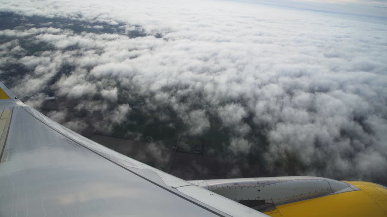 POV airplane passenger window: white blanket and billowing clouds below with view of aircraft wing and yellow engine in flight, handheld slow motion