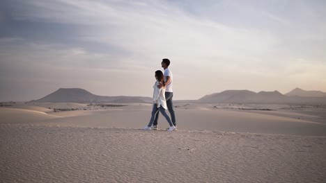 Slow-motion-footage-of-embraced-couple-walking-in-romantic-relationship-under-sun-and-blue-sky-in-desert.-Two-young-lovers-walking-by-desert-sand-in-casual-clothes.-Perfect-blurred-scene-of-mountains-on-the-background