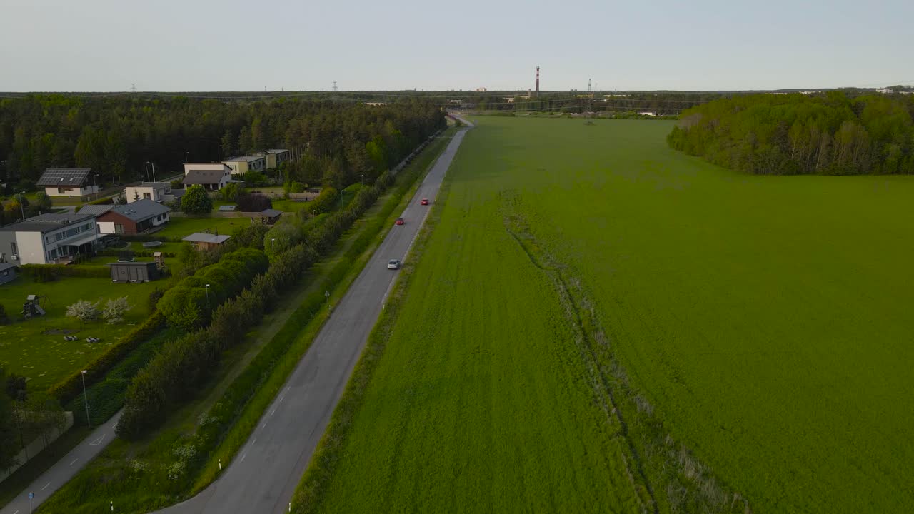 Beautiful aerial drone footage of small cars and vehicles driving on a rural countryside asphalt road surrounded by green grassy farm fields and small houses on the left side during a cloudy day.
