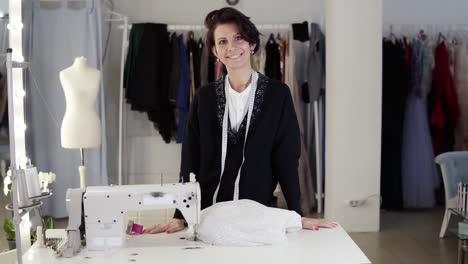 portrait of beautiful young short haired woman seamstress in elegant black sweater standing in workplace, touching measuring tape and looking at camera. dummy, garments, sketches and sewing machine are visible