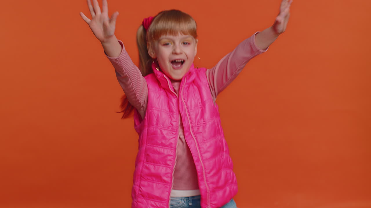 어린 소녀가 카메라를 향해 우호적으로 미소 짓고 손을 흔들며 인사 또는 작별 인사를 하고 환영합니다.