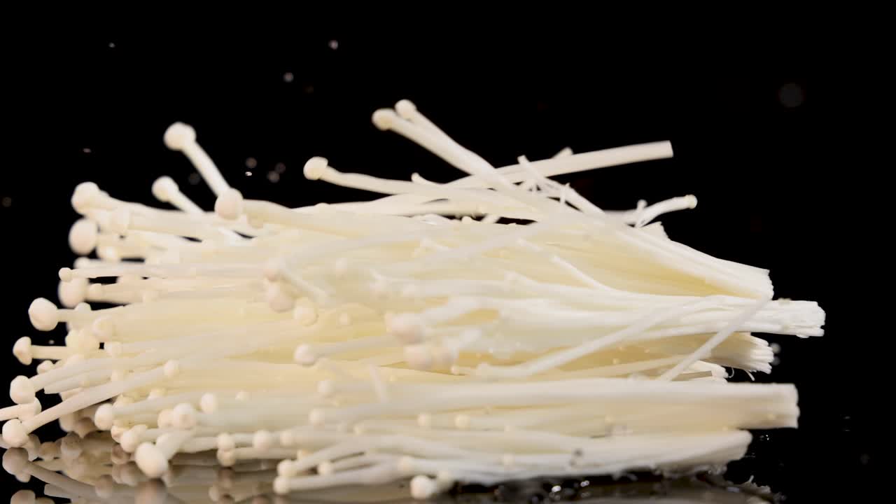 A cluster of enoki mushrooms falls and settles on a glossy black surface, captured in high-speed sequence with dramatic studio lighting and minimal background