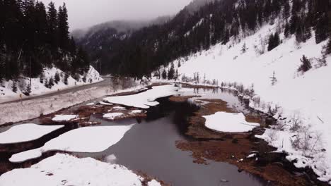 Die-Kamera-Bewegt-Sich-Entlang-Der-Straße-Und-Schneebedeckten-Bergen-Und-Fängt-Die-Spiegelungen-Des-Himmels-Im-Gefrorenen-Fluss-Ein