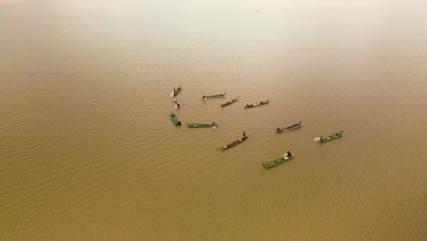 Multiple-fishermen-in-bright-colorful-boars-working-as-a-team-to-catch-a-shoal-of-fish-on-open-lake-in-Southeast-Asia,-birds-eye-drone-orbit