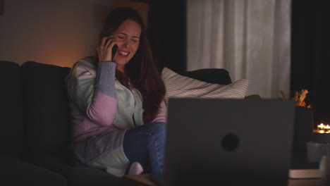 Mujer-Sonriente-Sentada-En-Un-Sofá-En-Casa-Por-La-Noche-Hablando-Por-Teléfono-Móvil-Y-Viendo-Una-Película-O-Un-Programa-En-Una-Computadora-Portátil