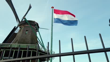 Dutch-red,-white-and-blue-flag-waving-in-the-wind-with-an-old-dutch-windmill-and-blue-sky-in-the-background-during-a-sunny-day