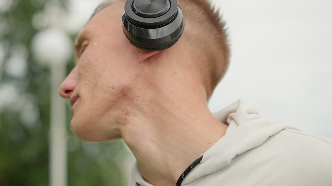 hombre blanco joven con auriculares, sudadera con capucha en el exterior, ajustando los auriculares, ojos cerrados, expresión pensativa, gotas de lluvia nubladas, horizonte nublado, primeros planos cinematográficos, banda sonora ambiental, estado de ánimo reflexivo, urbano