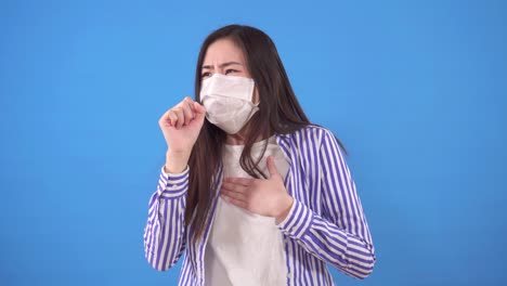 diseased beautiful young asian woman with a protective medical mask on her face coughs while standing on a blue background