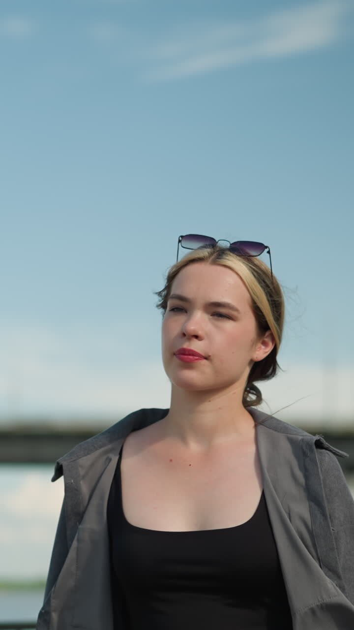 Woman in a black crop top and grey open shirt walks near a bridge, gazing into the distance, background features a blur of cars passing over the bridge under a clear blue sky