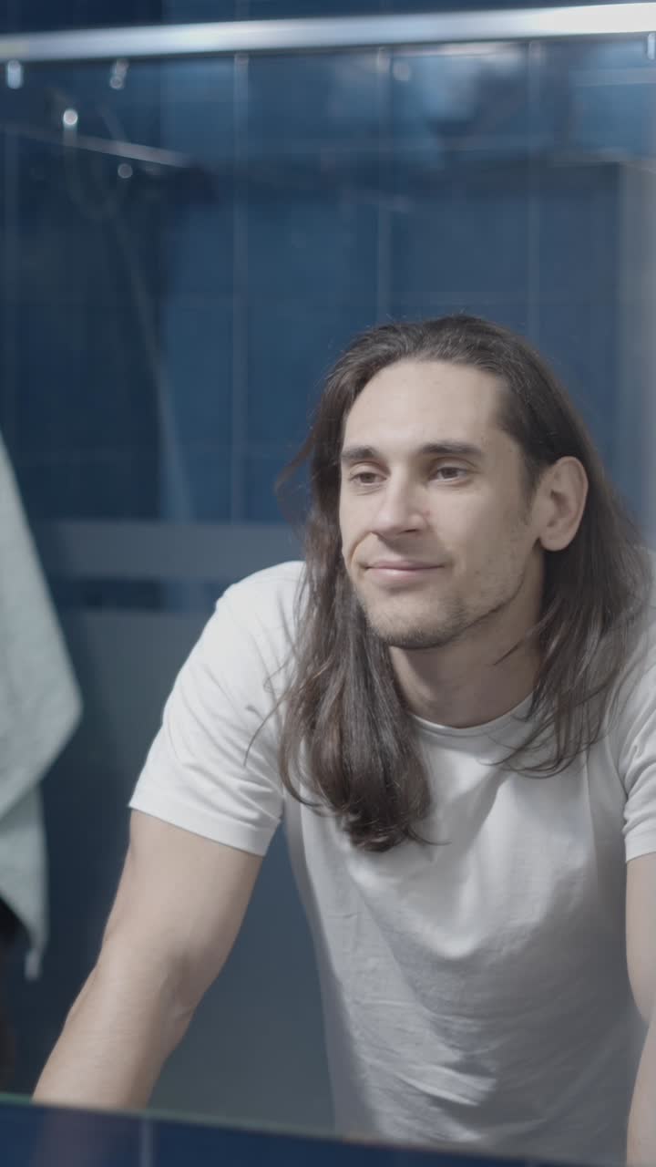 A man with long hair looking at his reflection in a bathroom mirror, showing various expressions