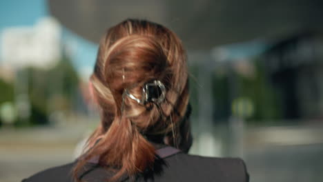 Close up rear view of professional woman in black suit with tied-back hair sipping coffee while on phone call walking near glass building with reflection of moving car in urban environment and soft sunlight