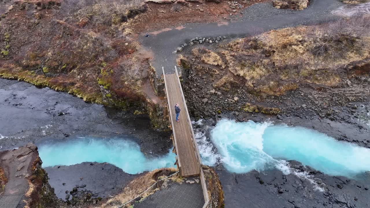 In the breathtaking beauty of Iceland's untamed landscape, a man gazes out from the precipice of a bridge, holding a drone in his grasp, Waterfall Bruararfoss