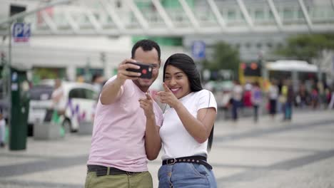 Feliz-Pareja-Joven-Tomando-Selfie-Con-Teléfono-Inteligente-En-La-Plaza