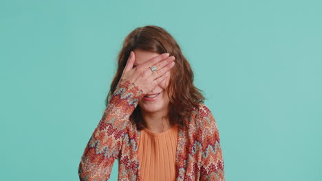 a beautiful woman is pointing at the camera with a surprised expression on her face. she is wearing a colorful jacket and a orange top. she is in front of a blue background.