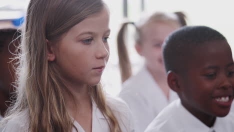 group of elementary pupils wearing uniform sitting on floor raise hands to answer question in class