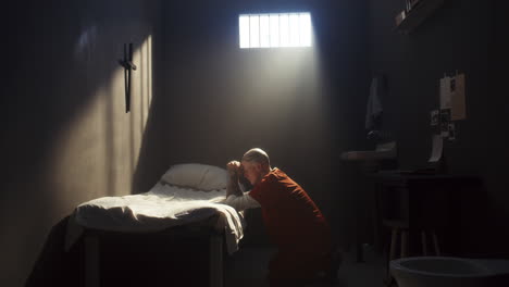 tattooed inmate kneeling by bed and praying during day in prison cell