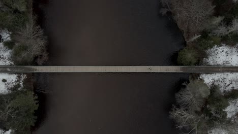 Man-Walking-Through-A-Narrow-Bridge-Over-The-Calm-River-In-Vallee-Bras-du-Nord-In-Quebec,-Canada---Aerial-Drone-Shot