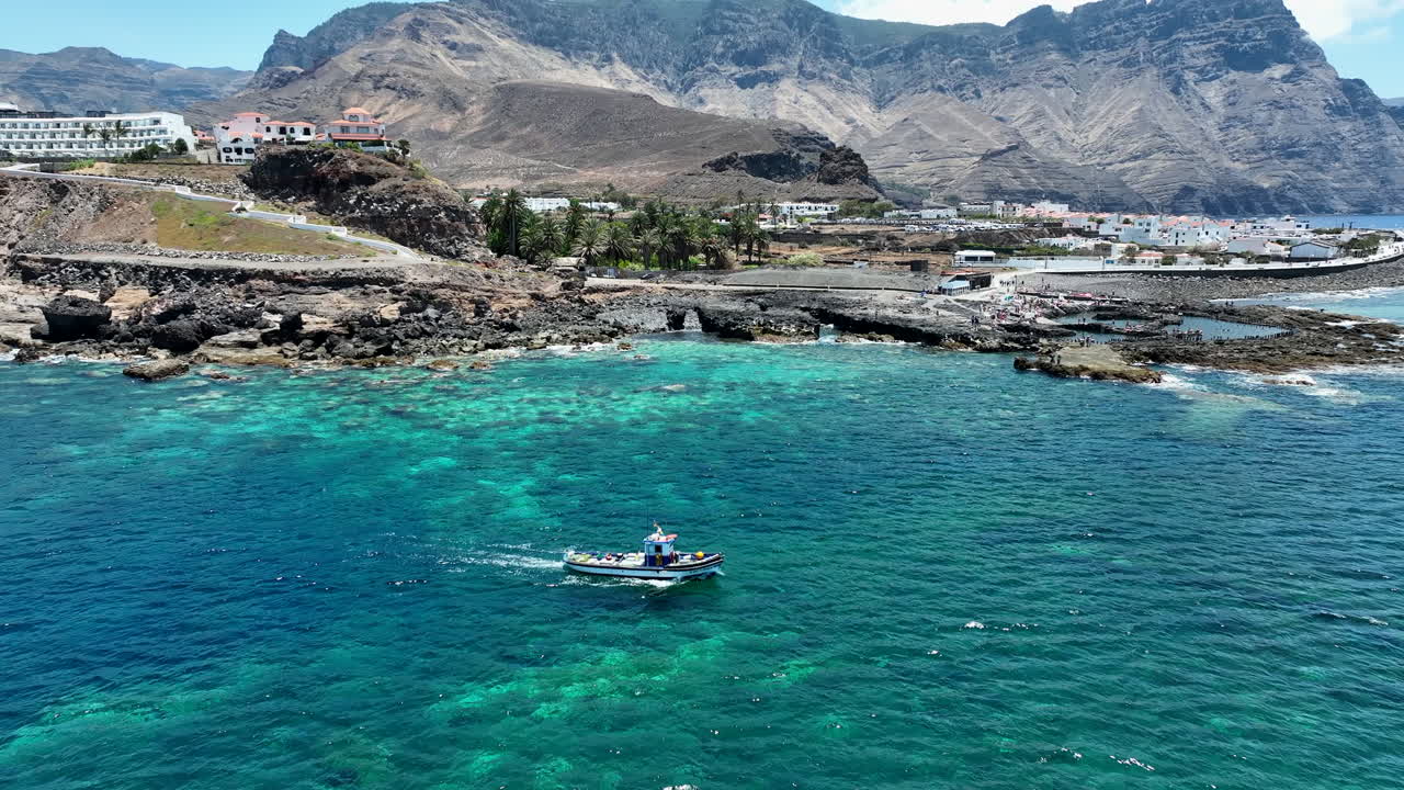maravillosa toma aérea siguiendo un barco de pesca en las aguas turquesas del puerto de agaete en un día soleado y donde se pueden ver las montañas y la costa