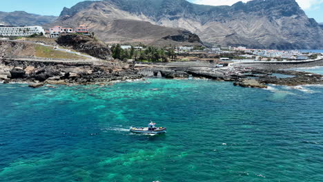 Wunderschöne-Luftaufnahme,-Die-Einem-Fischerboot-Im-Türkisfarbenen-Wasser-Des-Hafens-Von-Agaete-An-Einem-Sonnigen-Tag-Folgt-Und-Wo-Man-Die-Berge-Und-Die-Küste-Sehen-Kann