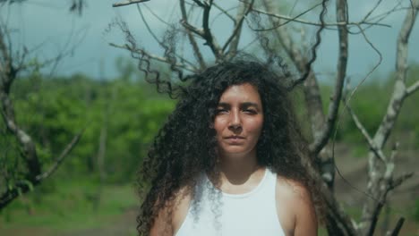 a woman with long curly dark hair and middle eastern features playfully sticks out her tongue while facing the camera, standing in front of sparse trees during a slow dolly-in shot, slow motion