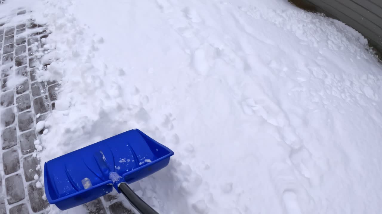 POV som skyver en blå spade langs kanten av en innkjørsel, fjerner nysnø og synlige fotspor til en pen brøytekant ved siden av en husvegg under vinterlig rydning av fortau