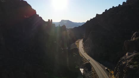 4k aerial view of a curvy mountain road with the sun high in the sky and cliffs on the side with lens flare and a creek running below