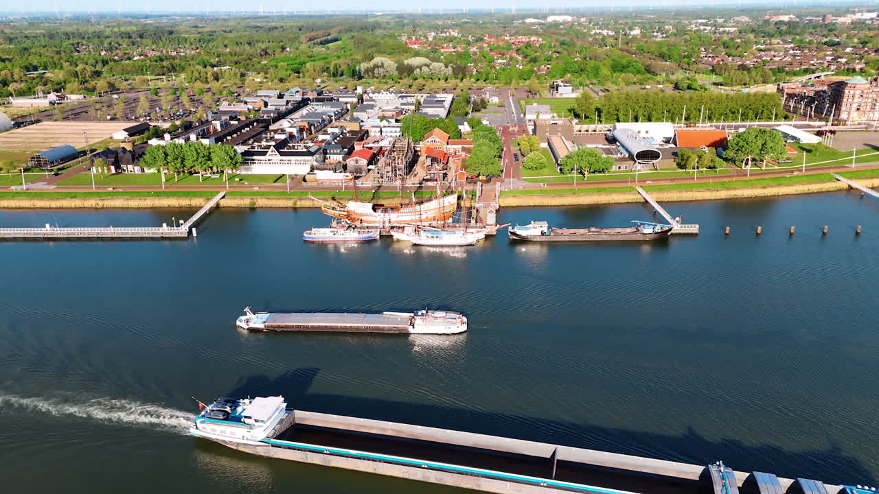 Distancing from the reconstruction of the ship Batavia and other boats around it. Barge moves by the waterscape. Cityscape of Lelystad, the Netherlands at backdrop.