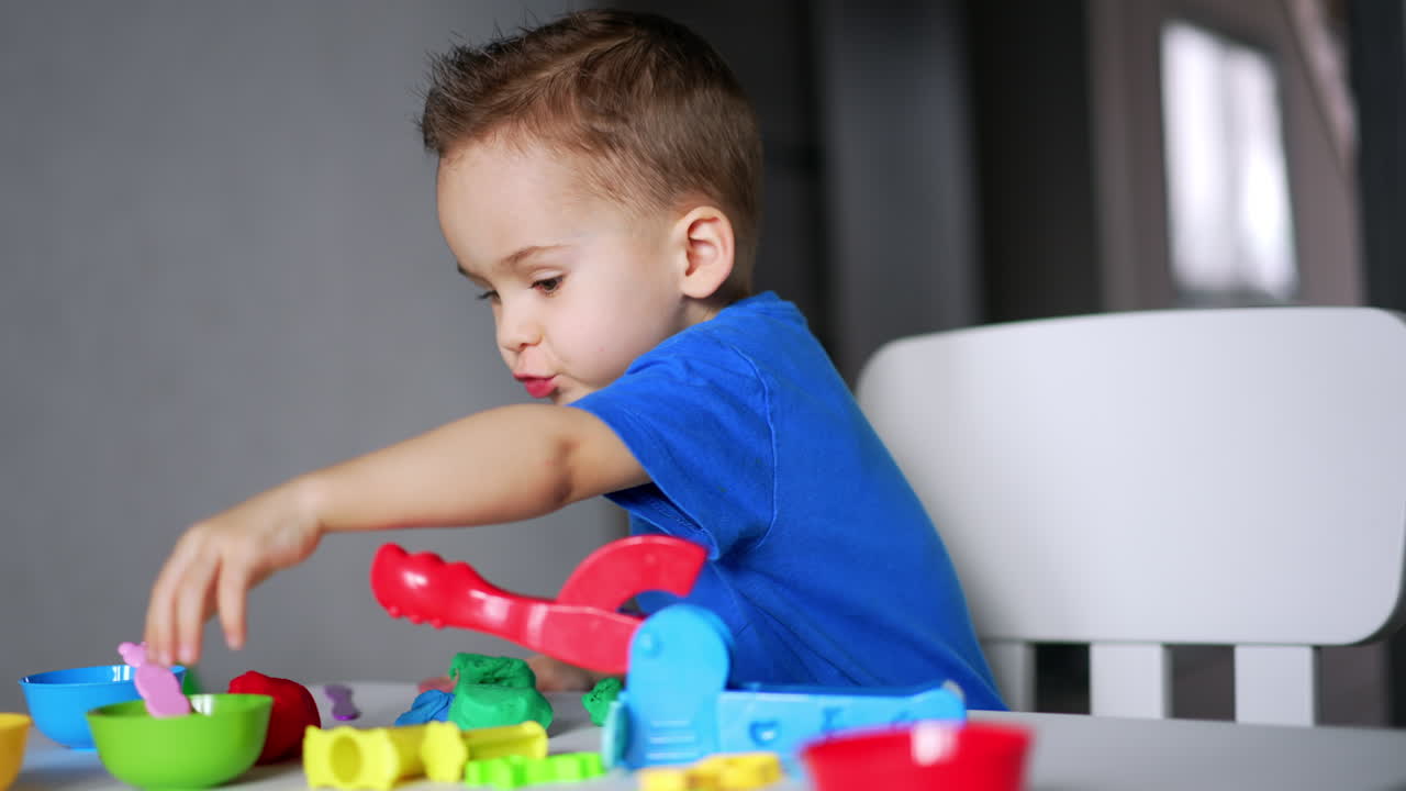 Concentrated Caucasian toddler sits at his desk at home. Cute baby boy sculpturing with plasticine.
