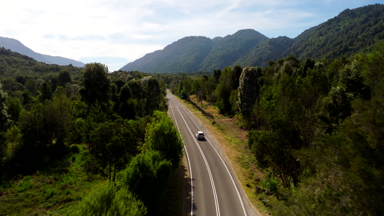 Un dron sigue a un coche por una carretera de dos carriles cerca del río Petrohué en Chile, serpenteando a través de un bosque exuberante y las estribaciones de las montañas bajo la suave luz del verano