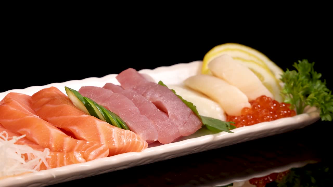 A pair of chopsticks selects salmon roe from an assorted sashimi sushi set on a white plate, under studio lighting with a black background