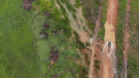 a vehicle and trailer drive across a dirt road stream, surrounded by lush greenery