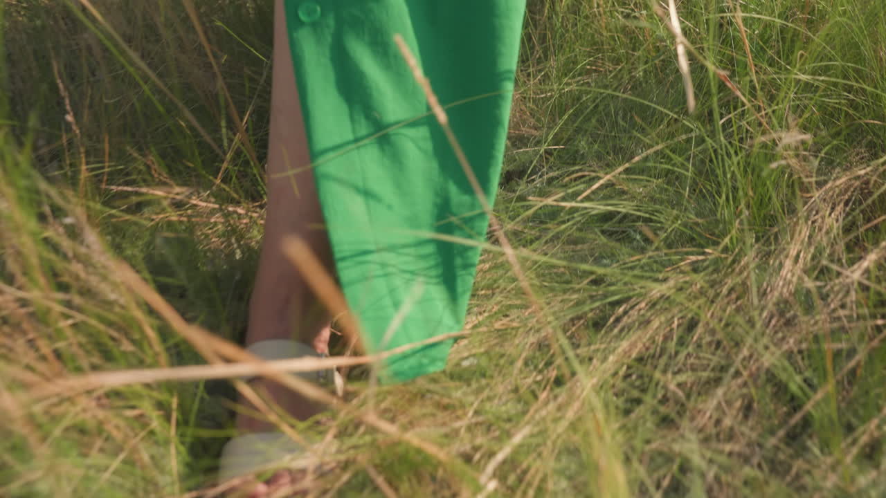lower angle view of lady foot in white sandal stepping through overgrown grassy field in green gown, blades parting as she moves gently forward under warm afternoon