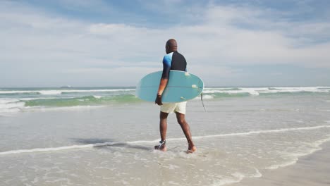 hombre mayor afroamericano caminando por una playa sosteniendo una tabla de surf y corriendo hacia el mar