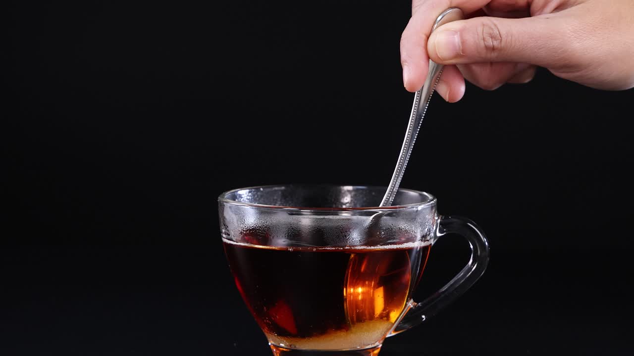 A hand stirs tea in a glass cup against a black background, highlighting the motion and simplicity of the action