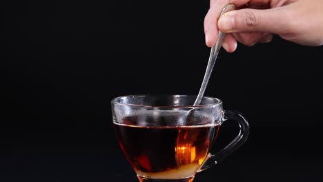 a hand stirs tea in a glass cup against a black background, highlighting the motion and simplicity of the action