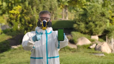 portrait young man in a protective suit, glasses, a respirator and gloves holds a smartphone with green screen and chroma key with vertical screen position and points at it with a finger. close up