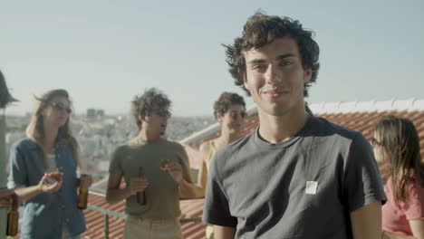 retrato de un joven caucásico mirando la cámara y sonriendo mientras tiene una fiesta en la azotea con amigos