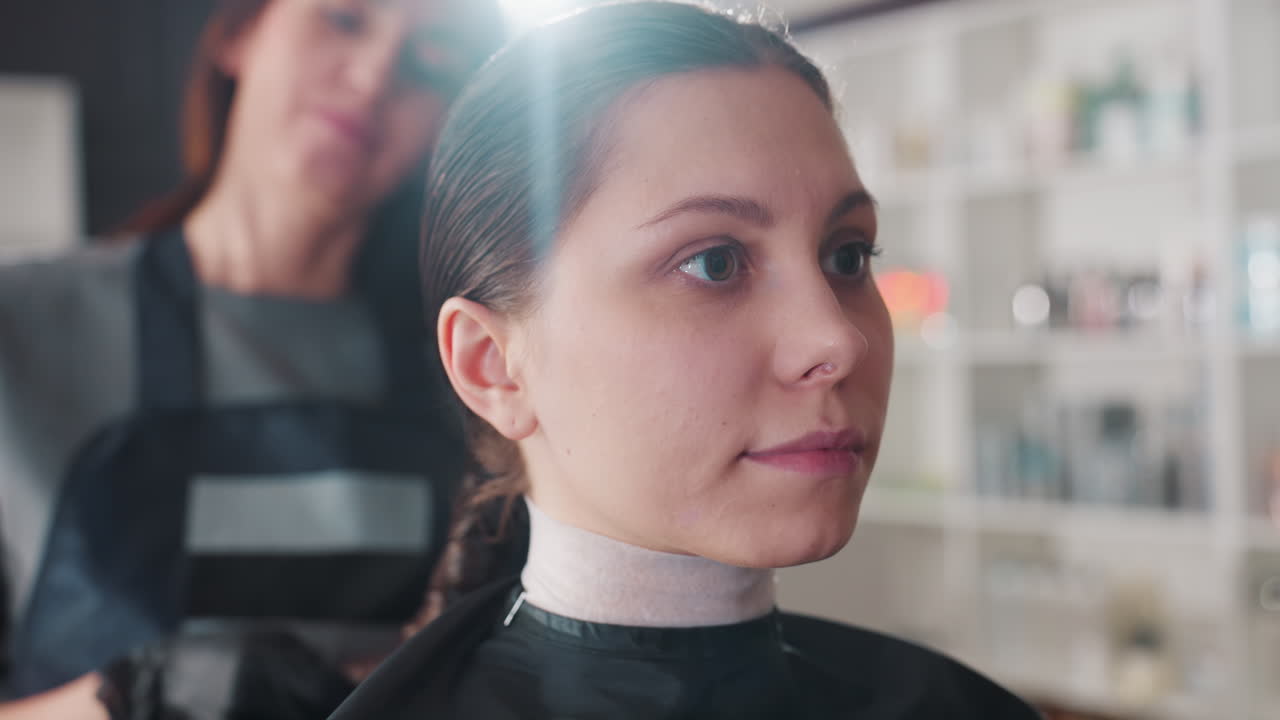 primer plano sereno de una clienta después de peinarse con un estilista difuminado detrás, mujer caucásica mostrando una expresión de calma y una línea de cabello cuidada, suave destello de flash de estudio, fondo de salón profesional y limpio