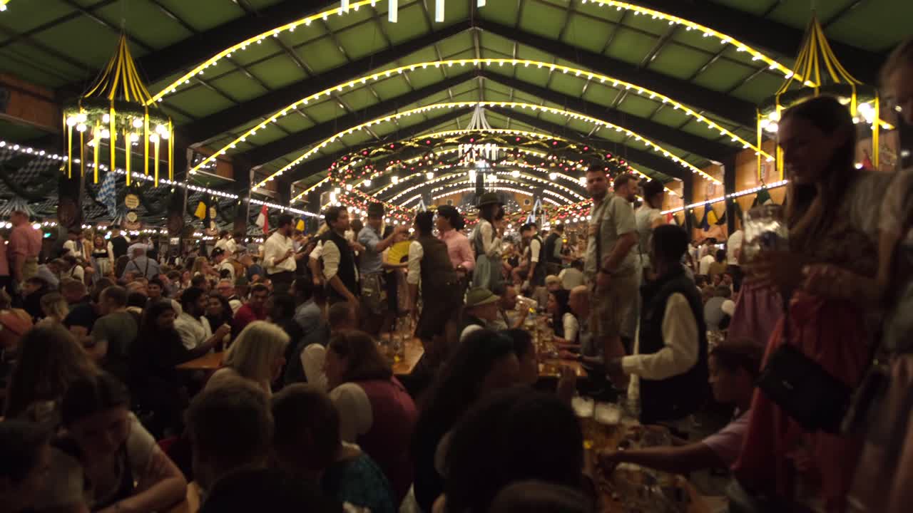 Men and women dance and drink inside a large Oktoberfest tent, celebrating with lively music, traditional costumes, and joy.