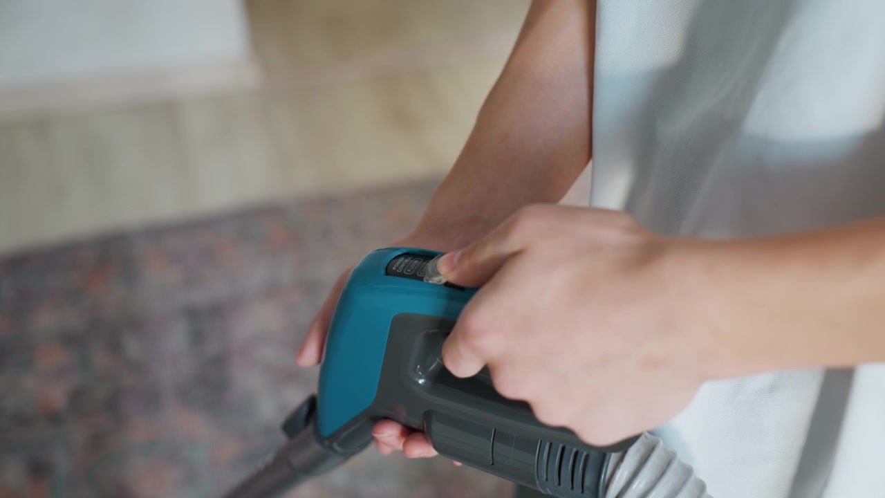 Gros plan des mains manipulant la poignée d'un aspirateur montrant le tuyau et le bouton, aspiration douce sur un tapis à motifs dans le salon, lumière naturelle du jour, tâche ménagère concentrée et entretien soigné de la maison avec ergonomie.