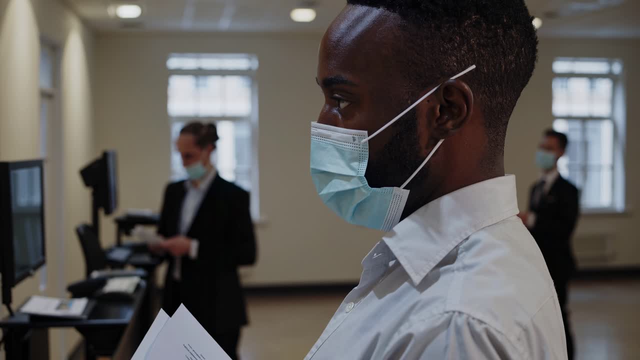 Professional businessman wearing protective face mask analyzing document while colleagues working on computers in modern corporate workplace