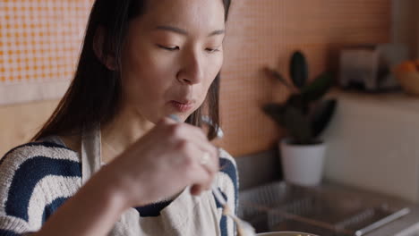 joven asiática desayunando en la cocina disfrutando de cereales de avena saludables mezclados con fruta relajándose en casa el fin de semana