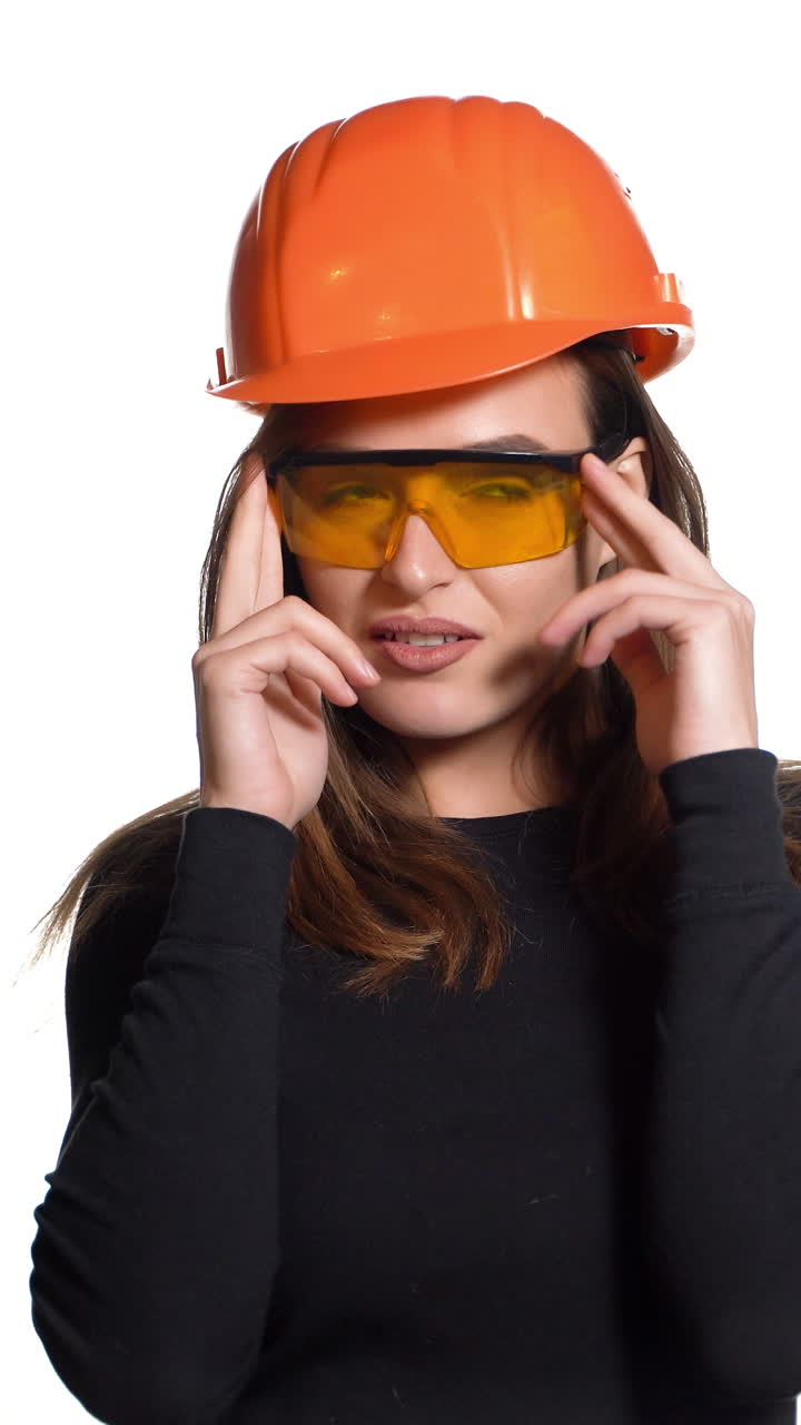 A joyful woman in orange helmet for building is trying on goggles and posing in it on a white background in the studio. Vertical video