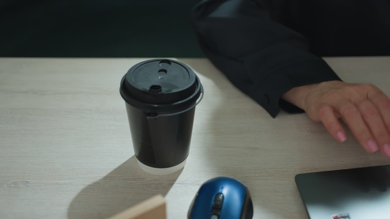 Top down view of person placing takeaway tea cup on wooden desk near computer mouse and real estate miniature model, preparing workspace for productive work session in bright modern office