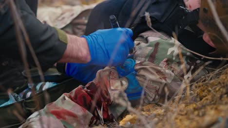 cerca un médico hombre confiado con ropa verde oscuro y guantes azules escribe el grupo sanguíneo en la ropa de un soldado herido que yace en el suelo mientras está herido y proporciona primeros auxilios durante las operaciones de combate