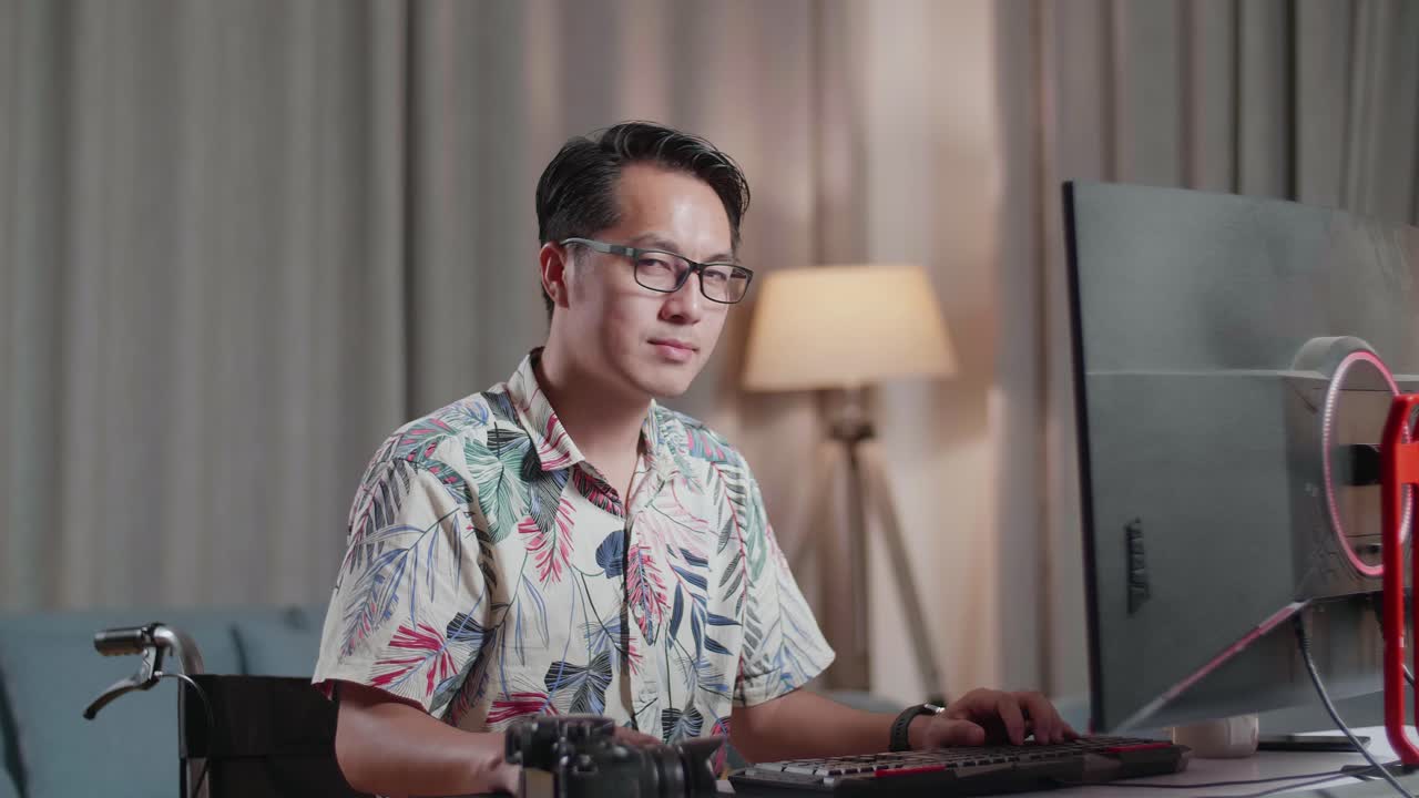 체어 에 있는 아시아인 남자 가 집 에 있는 카메라  에 있는 데스크 으로 비디오 를 편집 하는 동안 카메라 에 미소 짓고 있다