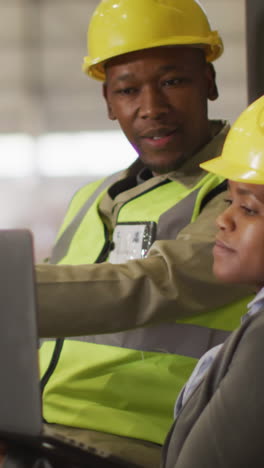 african american male and female workers wearing safety suits and using laptop in warehouse