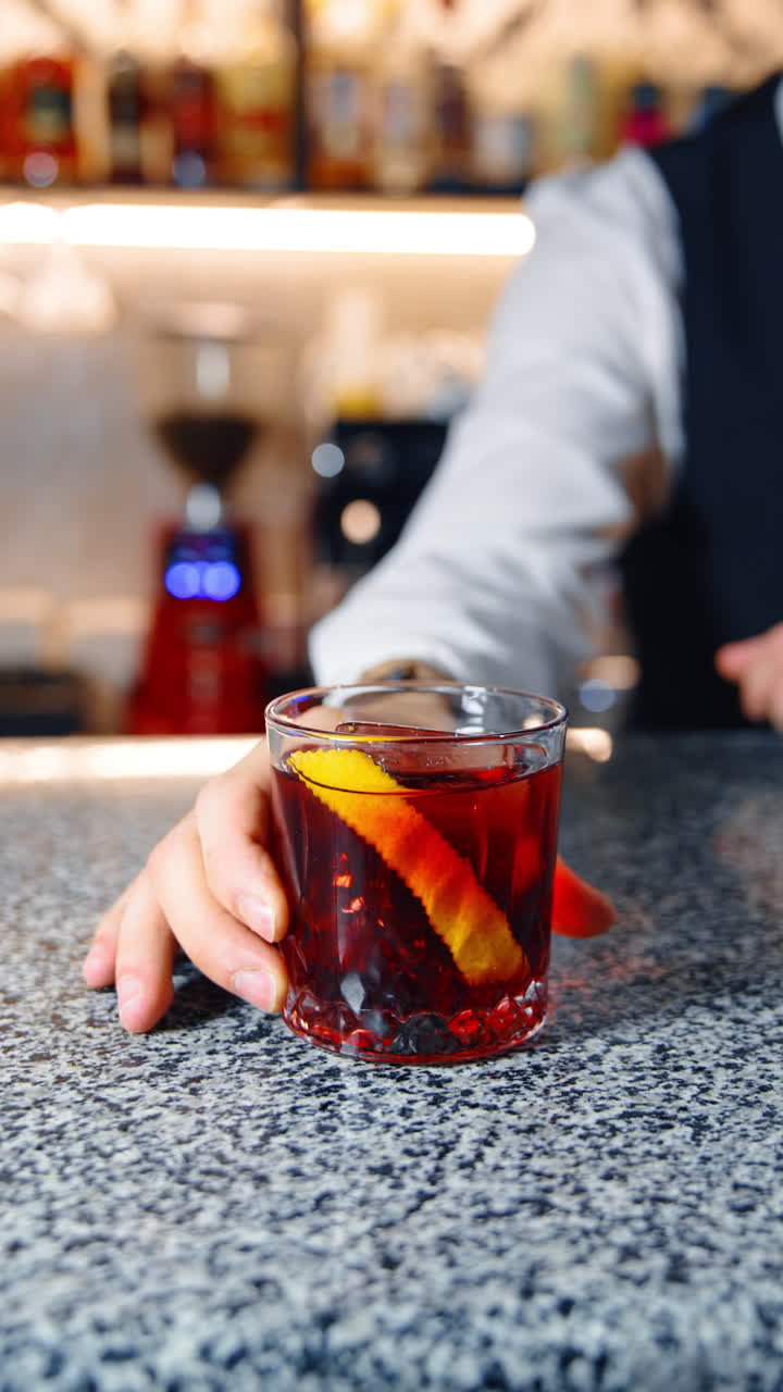 Male hand of unrecognized bartender wearing a white shirt and black vest holding a glass with brown drink. Beverage with a lemon slice and ice cube in the bar. Vertical video.